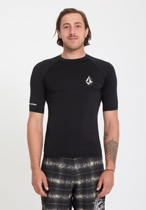 Homme aux cheveux mi-longs portant un rashguard noir à manches courtes et un short de bain à carreaux noirs, debout devant un fond blanc uni.