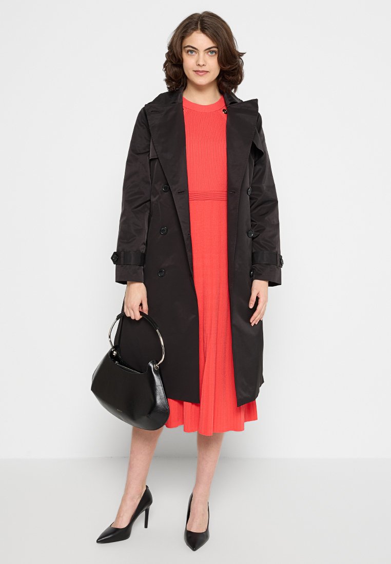 Czarny płaszcz trench z podwójnymi guzikami, noszony na koralowej sukience z plisami. Czarna torba i czarne szpilki dopełniają look.