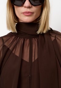 Bruine doorschijnende blouse met een hoge halslijn en een vloeiende uitstraling met geplooide accenten. De stof lijkt zacht en lichtgewicht te zijn.