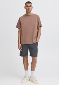 Lysebrun bomulds-t-shirt med en afslappet pasform, kombineret med mørkegrå bomuldsshorts. Modellen har hvide sokker og cremefarvede sneakers på. Simpelt design.