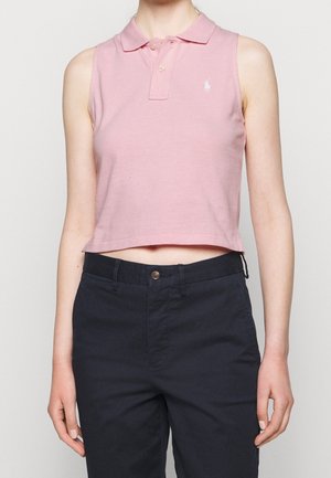 Kobieta ubrana w różowy, bezrękawnikowy, krótki polo oraz ciemnogranatowe spodnie, stoi na tle jasnego, jednolitego tła.