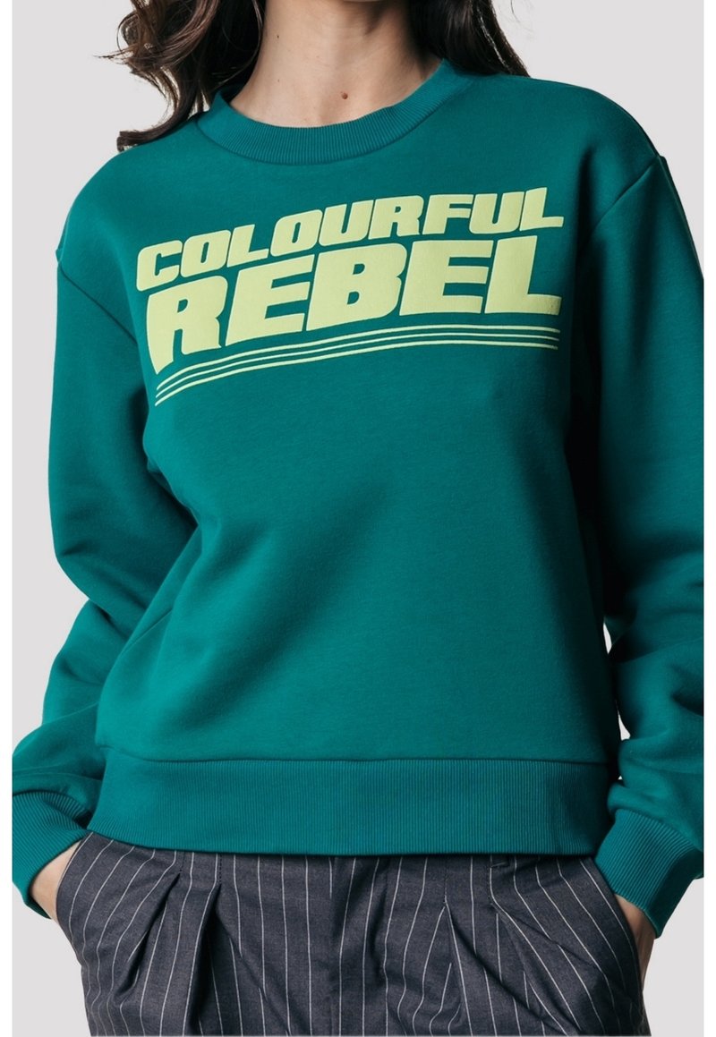 Felpa turchese con il bold di testo giallo chiaro "RIBELLE COLORATO" sopra tre linee orizzontali, indossata con pantaloni scuri a righe e mani nelle tasche.