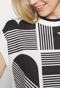 Short-sleeve top in white cotton featuring bold black graphic patterns, including lines and shapes, with a crew neckline and relaxed fit.