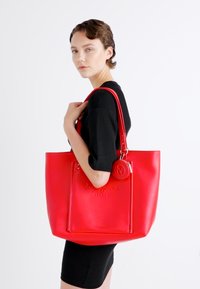 Borsa tote in pelle sintetica rossa con accento del logo, tasche con zip e doppi manici. Texture liscia e design strutturato.