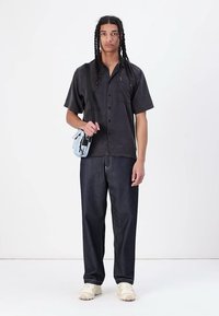 Schwarzes Kurzarmhemd, blaue Umhängetasche, dunkle Jeans und helle Turnschuhe. Das Hemd hat eine durchgehende Knopfleiste und einen lockeren Schnitt.