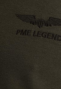 Teksturna tkanina maslinaste boje s urezanim logotipom s krilima i natpisom "PME LEGEND." Bez dodatnih uzoraka ili akcenta.