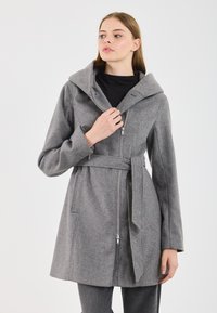 Donna che indossa un cappotto grigio con cintura, dotato di un grande colletto e zip, tiene il colletto con una mano su uno sfondo bianco semplice.
