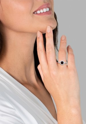 Zilveren ring met een vierkante zwarte steen, omringd door kleine heldere stenen. De hand van het model rust nabij haar gezicht, waardoor het ontwerp van de ring goed zichtbaar is.