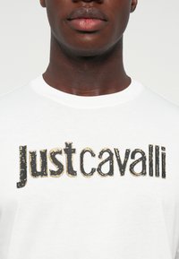 Tuvplāns uz cilvēku, kurš valkā baltu T-kreklu ar melniem un zeltīti nolietotiem burtiem ar uzrakstu "just cavalli" uz krūšu.