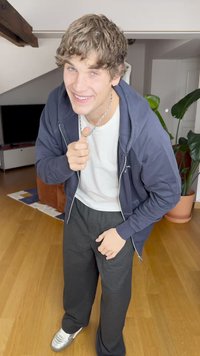 Model trägt einen navyfarbenen Reißverschluss-Hoodie, ein weißes T-Shirt und schwarze lässige Hosen. Sie trägt silberne Sneaker und eine Kettenhalskette und steht in einem Innenraum.