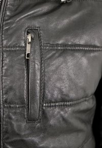 Schwarze Lederjacke mit einer Reißverschlusstasche, gesteppter Struktur und präzisen Nahtdetails entlang der Nähte.