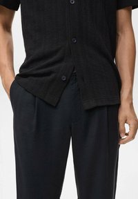 Chemise noire à manches courtes texturée avec fermeture à boutons, associée à un pantalon sombre. La chemise présente une coupe décontractée et un motif de tissage subtil.