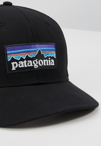 Căciulă de baseball neagră cu o margine curbată. Prezintă o insignă brodată rectangulară cu un design de munte în nuanțe de albastru, portocaliu și mov și text.