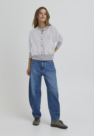 Light gray cropped sweatshirt with button closure, ribbed cuffs, and logo patch on chest, paired with loose blue jeans and gray sneakers.