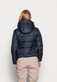 Marinblå puffjacka med huva, quiltat mönster och ribbade muddar. Bärs över ljusbeige byxor i sweatmaterial med en jämn, glänsande yta.