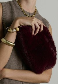 Bordeaux kleurige nep-fur clutch met een zachte textuur, ronde vorm en een pluche afwerking, gehouden naast een gouden kettingarmband en een bewerkte polsaccessoire.