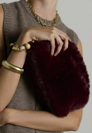 Bordeaux kleurige nep-fur clutch met een zachte textuur, ronde vorm en een pluche afwerking, gehouden naast een gouden kettingarmband en een bewerkte polsaccessoire.