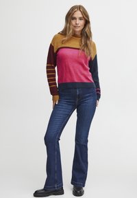 Pull en tricot à col montant, avec des rayures orange, rose et bleu marine ; associé à un jean bleu évasé et des bottes noires.