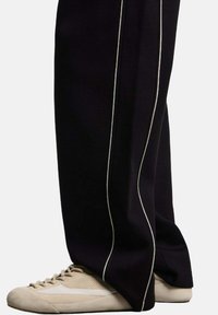 Zwarte wijde broek met witte zijstrepen, gemaakt van een textuurstof. Gecombineerd met beige low-top sneakers met een mix van materialen.