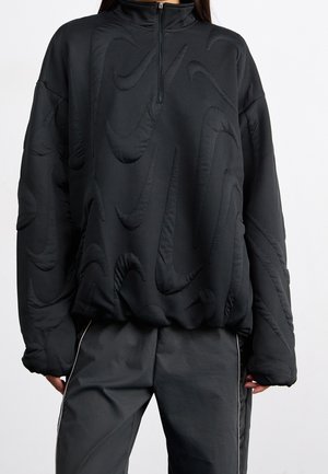 Persoon met een zwarte trui met reliëf swoosh-patronen en zwarte broek met witte zijstrepen, getoond van schouders tot halverwege het dijbeen.