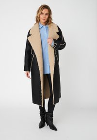 Svart lang frakk med beige lammeskinnskrage og fôr, med kontrasterende sømmer, brukt over en lyseblå rutet skjorte og svarte bukser.