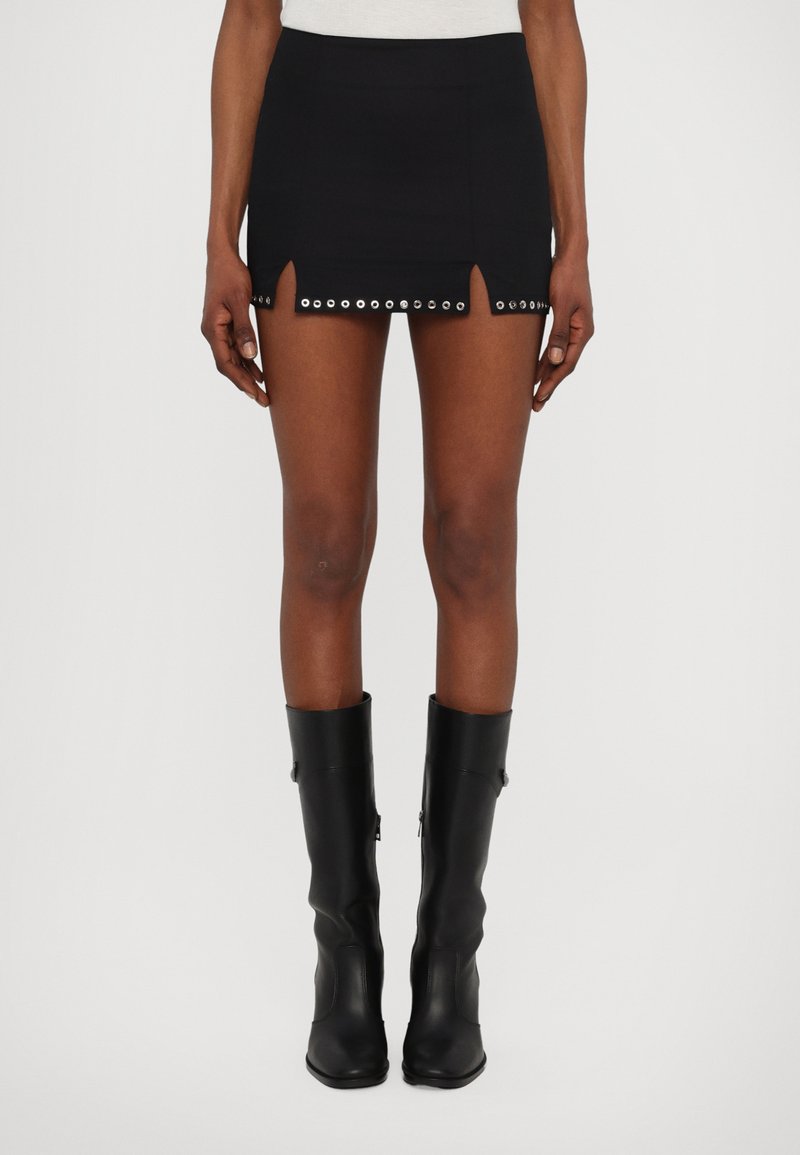 Μαύρη μίνι φούστα με κοπή στο στρίφωμα, διακοσμημένη με κυκλικούς μεταλλικούς οπλισμούς. Το υλικό φαίνεται λείο και εφαρμοστό.