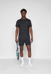 Svart poloskjorte med hvit kant, parret med svarte shorts og hvite joggesko. Holder en tenniskølle, står mot en nøytral bakgrunn.