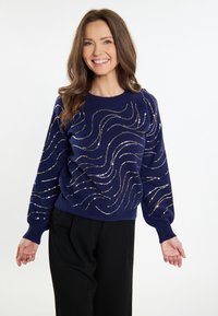 Navyblauer Pullover mit silbernen und goldenen Wellenmustern, gestrickter Stoff, runder Ausschnitt und weit geschnittene Ärmel. In Kombination mit schwarzen Hosen.