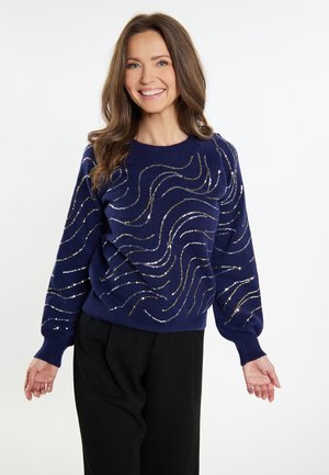 Navyblauer Pullover mit silbernen und goldenen Wellenmustern, gestrickter Stoff, runder Ausschnitt und weit geschnittene Ärmel. In Kombination mit schwarzen Hosen.