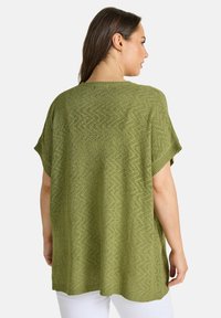 Mujer vistiendo un top de punto de manga corta texturizado en verde oliva, visto desde atrás, combinado con pantalones blancos sobre un fondo liso.
