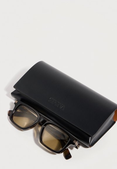 Μαύρα ορθογώνια γυαλιά ηλίου με φακούς καφέ απόχρωσης, τοποθετημένα κάτω από μια μαύρη δερμάτινη θήκη με ανάγλυφο το λογότυπο "Zegna" πάνω σε λευκή επιφάνεια.