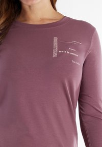 Woman wearing mauve long-sleeve shirt with text on chest reading "WELLBEING, practice yoga, be grateful, walk in nature, Tom Tailor."