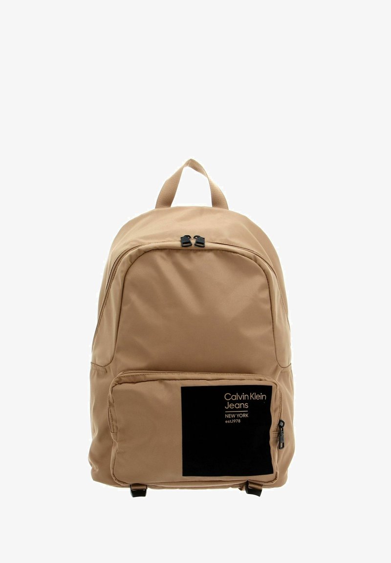 Sac à dos beige avec une texture lisse, double fermeture éclair, une poche avant avec un patch logo noir, et une poignée supérieure pour le transport.