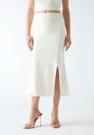 Femme portant une jupe midi blanche côtelée avec une fente à l'avant et des sandales à talons beiges, montrée de la taille aux pieds.