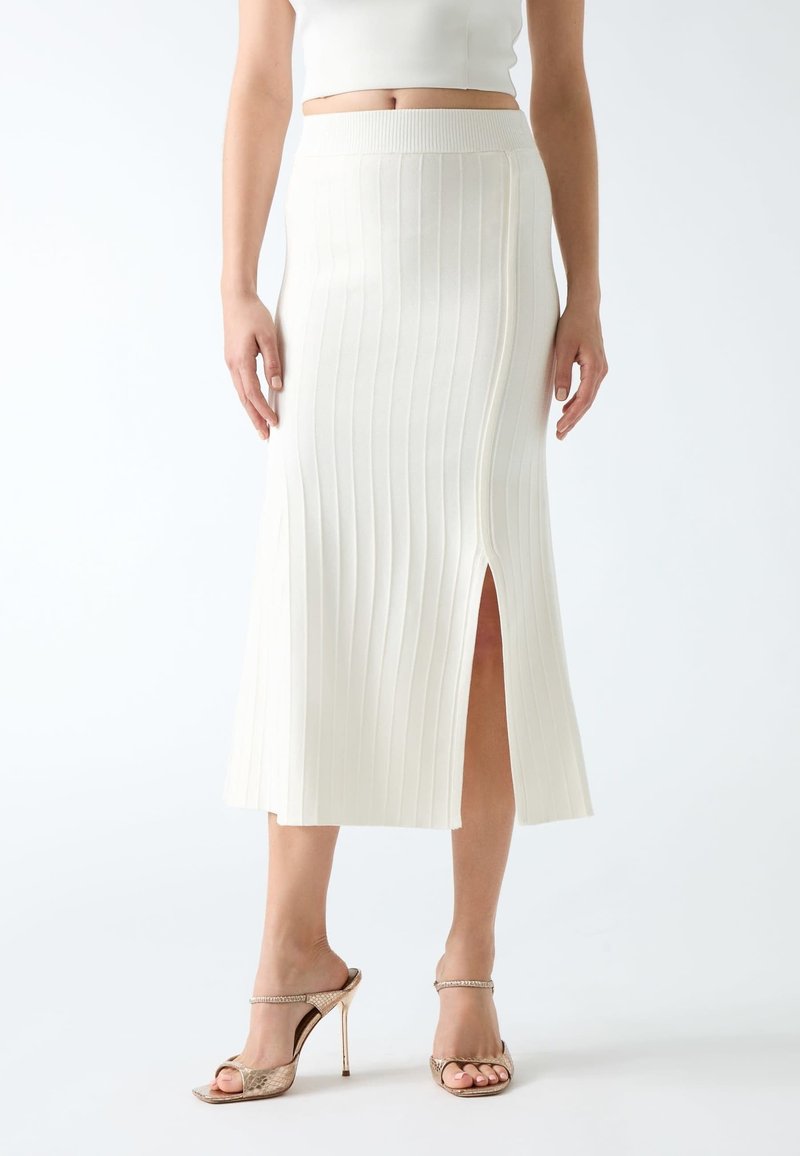 Femme portant une jupe midi blanche côtelée avec une fente à l'avant et des sandales à talons beiges, montrée de la taille aux pieds.