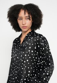 Fekete szatén blúz fehér pöttyökkel, klasszikus gallérral, gombos elejjel és hosszú ujjakkal, sima, fényes textúrával.