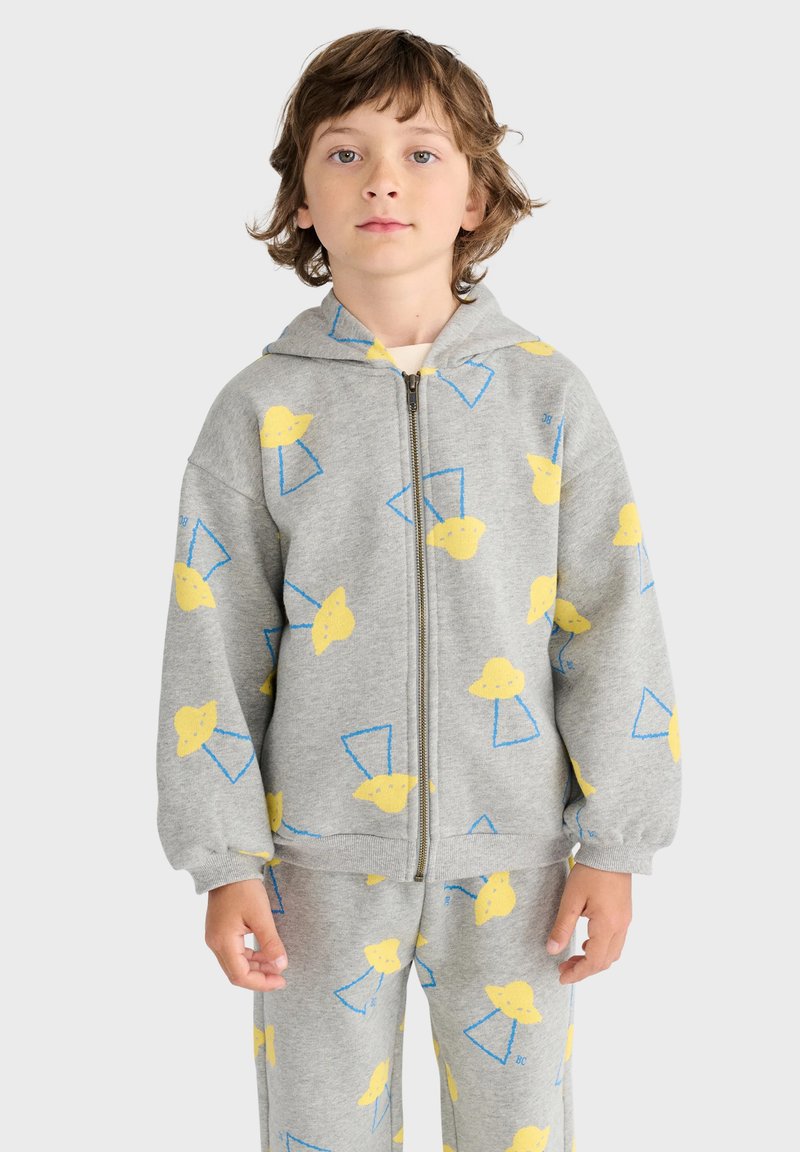 Enfant portant un sweat à capuche gris zippé et un pantalon avec des formes abstraites jaunes et bleues, debout devant un fond clair uni.