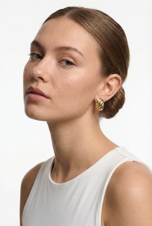 Jonge vrouw met lichtbruin haar in een staart, draagt een wit mouwloos topje en gedraaide gouden creolen, tegen een effen witte achtergrond.