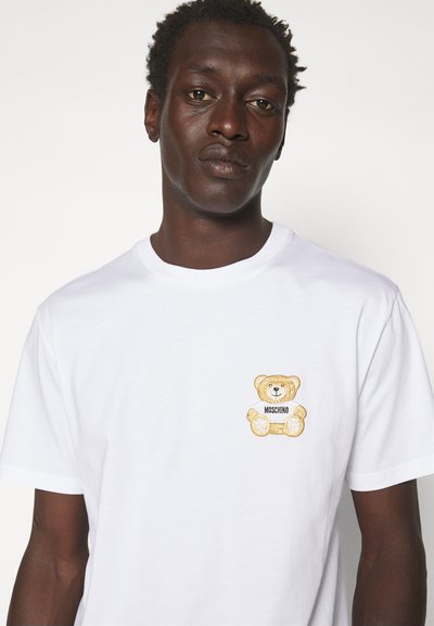 Camiseta de algodón blanca con un logo de oso de peluche bordado en oro y marrón en el lado izquierdo del pecho. Cuello redondo clásico y mangas cortas.