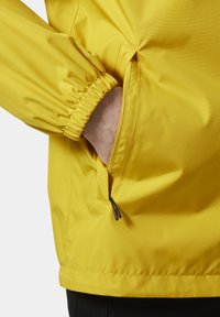 Chaqueta amarilla con puños elásticos y bolsillo lateral. Fabricada en un material liso, impermeable, con un acabado texturizado y cremallera negra en contraste.