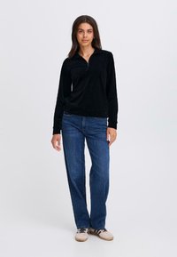Zwart corduroy top met een kwart-rits kraag, gecombineerd met blauwe denim jeans en beige sneakers met donkere strepen, staande tegen een neutrale achtergrond.
