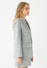 Blazer grigio a quadri con vestibilità su misura, davanti doppiopetto, rever a varco e due tasche frontali. Tessuto texturizzato con dettagli gialli.