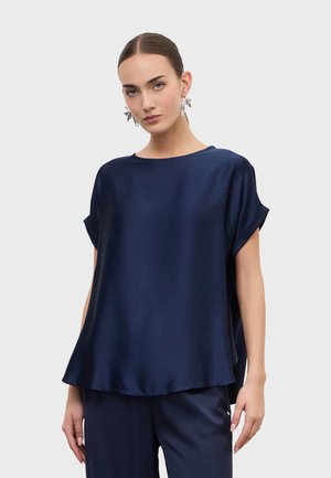Femme portant un chemisier ample en satin bleu marine avec des manches courtes retroussées et un pantalon assorti bleu marine, sur un fond clair uni.