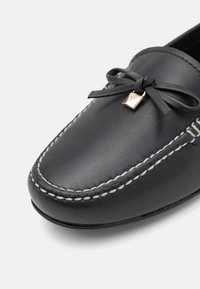 Mocassino in pelle nera con punta arrotondata, cuciture bianche e un dettaglio a fiocco. Presenta un piccolo ornamento in hardware dorato.