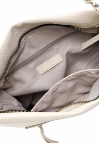 Cream-colored leather tote bag with a textured exterior, featuring a beige fabric interior, zippered pocket, and brand label inside.