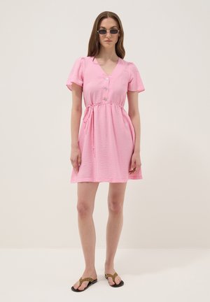 Femme en robe rose à manches courtes, boutonnée, avec ceinture nouée à la taille, portant des lunettes de soleil et des sandales plates marron, debout devant un fond clair uni.