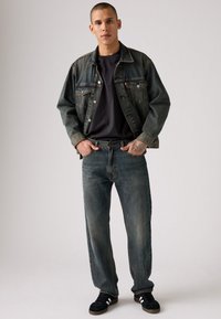 Chaqueta de mezclilla, azul oscuro con desgaste ligero, combinada con una camiseta negra y jeans rectos azules desvanecidos. Zapatillas negras completan el look.