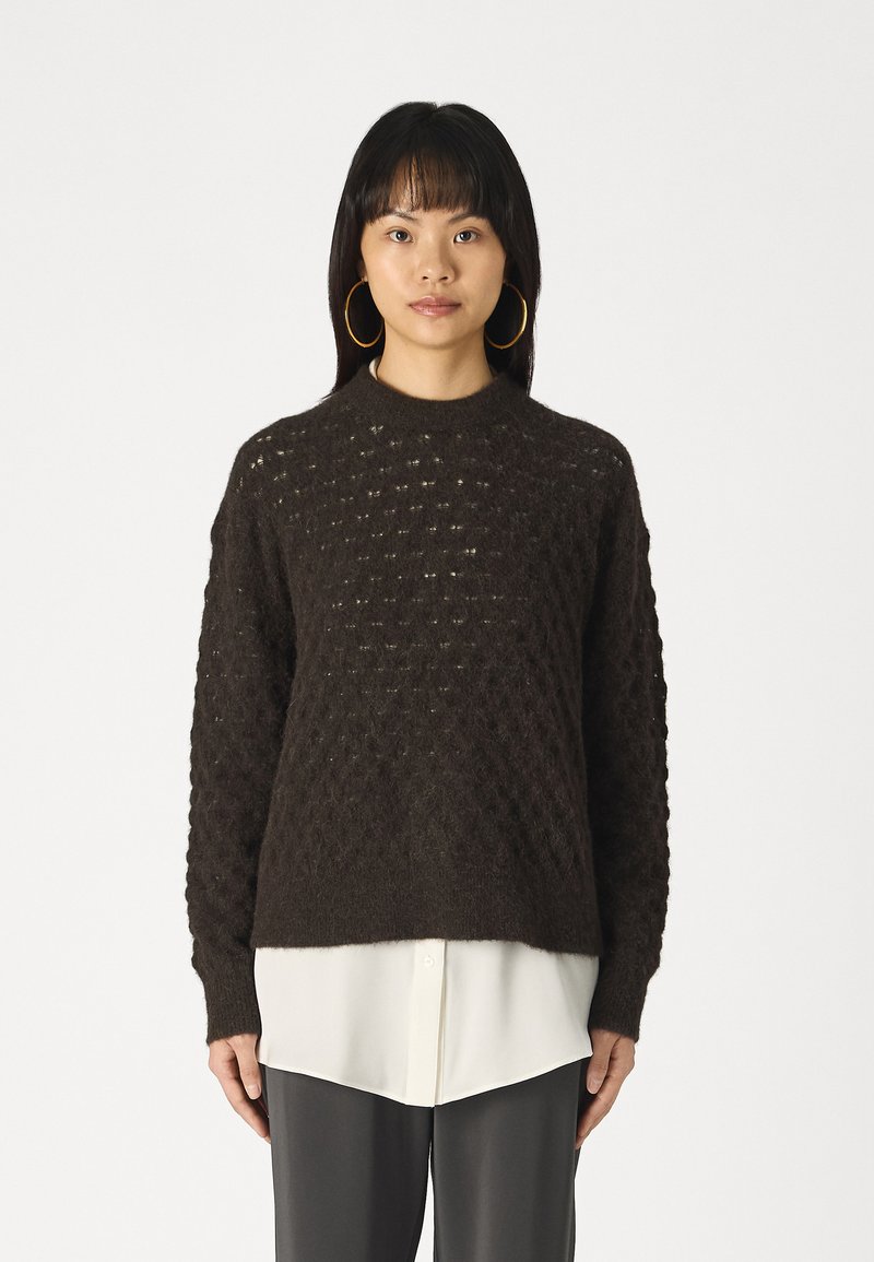 Pull en tricot marron foncé avec un motif en losanges texturé, col rond et coupe décontractée, superposé sur une chemise blanche et un pantalon gris.