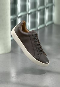 Brun sneakers i läder med textur, svarta skosnören och en vit gummisula. Har en minimalistisk design och en liten logotyp på sidan.