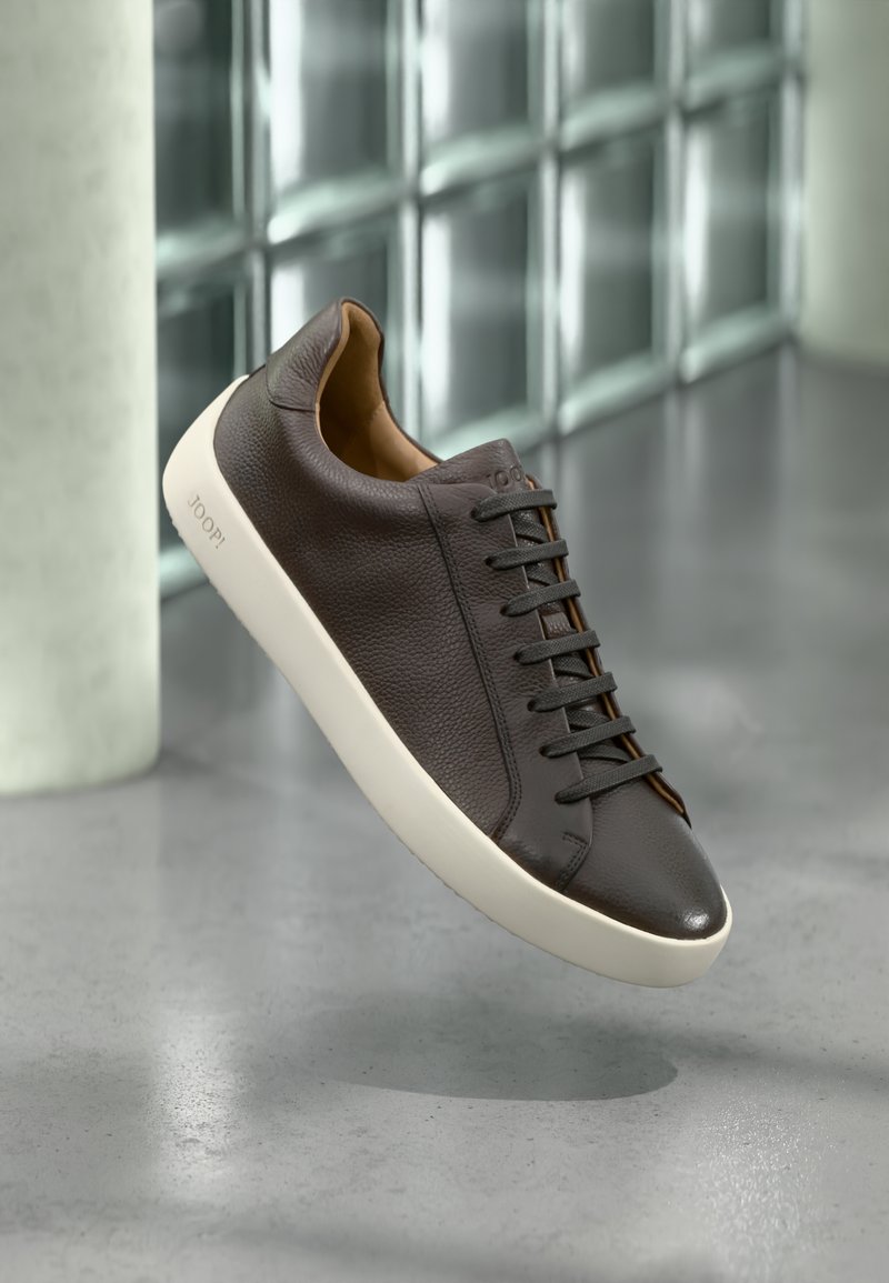 Brun sneakers i läder med textur, svarta skosnören och en vit gummisula. Har en minimalistisk design och en liten logotyp på sidan.